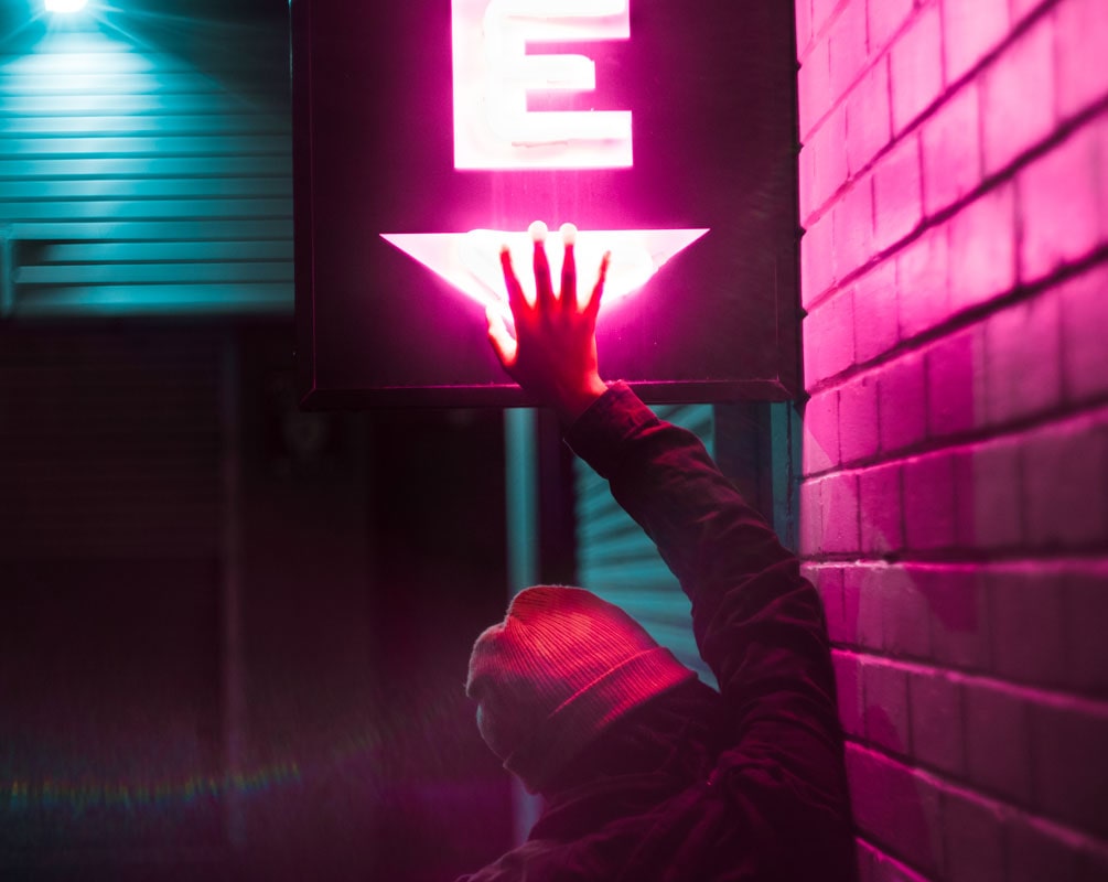 Back view of a person in a beanie with hand raised covering pink neon light sign symbolising digital life & it’s activation